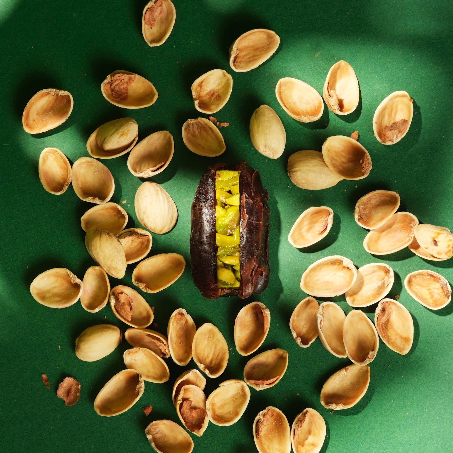 A top-down view of a single What a Date dark, hand-filled and hand-pitted organic dark date, sliced to reveal its generous filling of Sicilian pistachio, surrounded by the empty shells of the pistachios used in the filling, on a pistachio-green background.