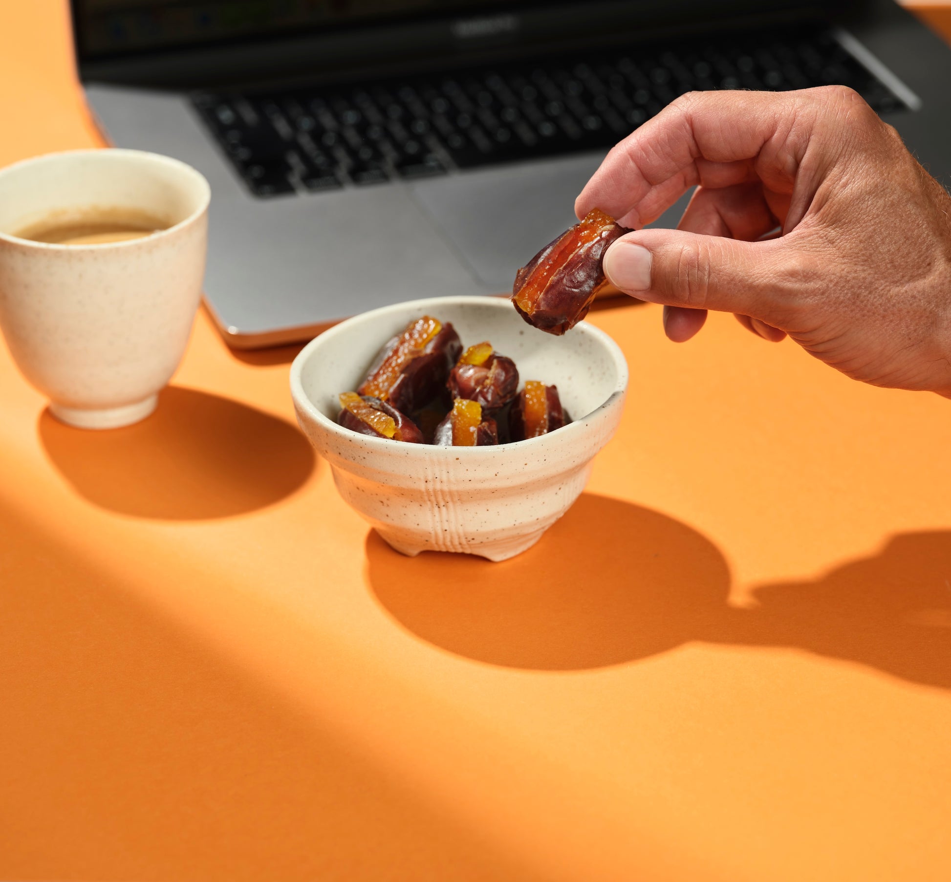 Person holding a date with orange peel from What a Date next to a bowl of dates and a cup of tea on an orange surface with a laptop in the background.