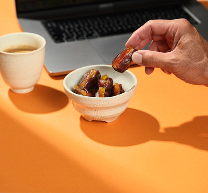 Person holding a date with orange peel from What a Date next to a bowl of dates and a cup of tea on an orange surface with a laptop in the background.