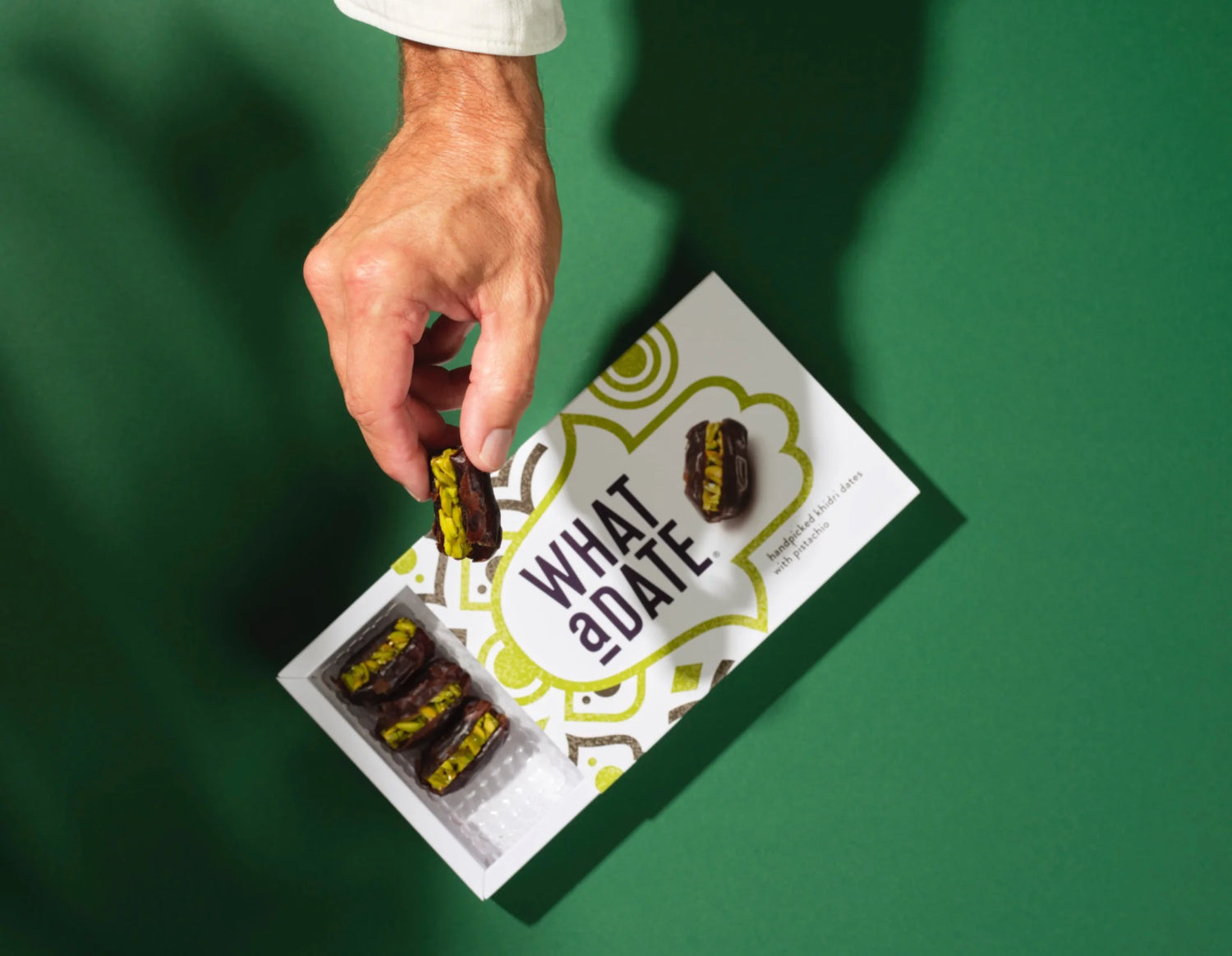 A hand picking up a single hand-filled What a Date Pistachio from its branded large box (12 pcs), on a pistachio-green background. The handmade filling of thick slivers of Sicilian pistachio gently baked in artisanal date syrup. 